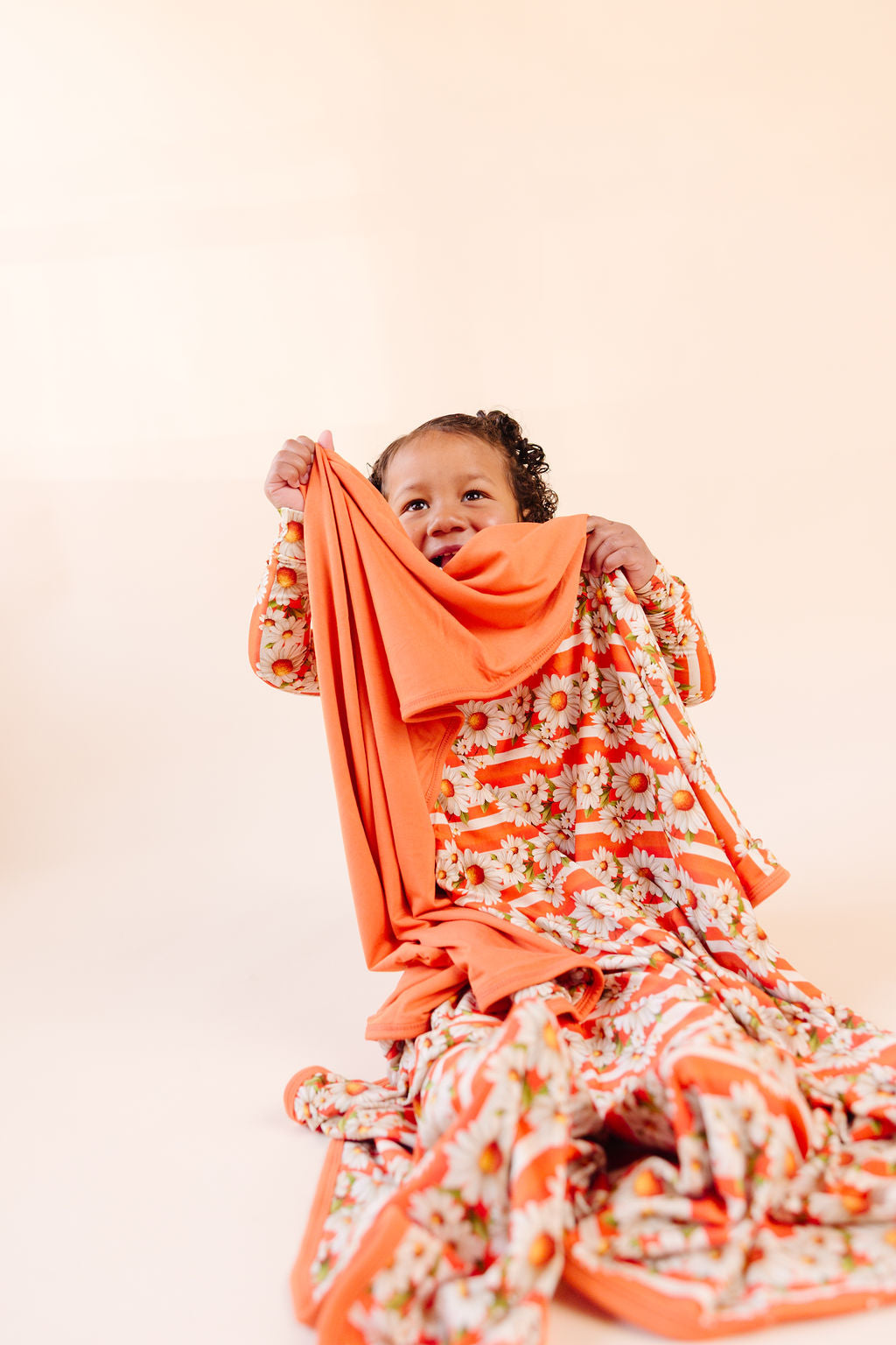 Child holding a bamboo reversible blanket orange and white stripe, with white daisy's on top  and the reverse side a bright orange against a beige background