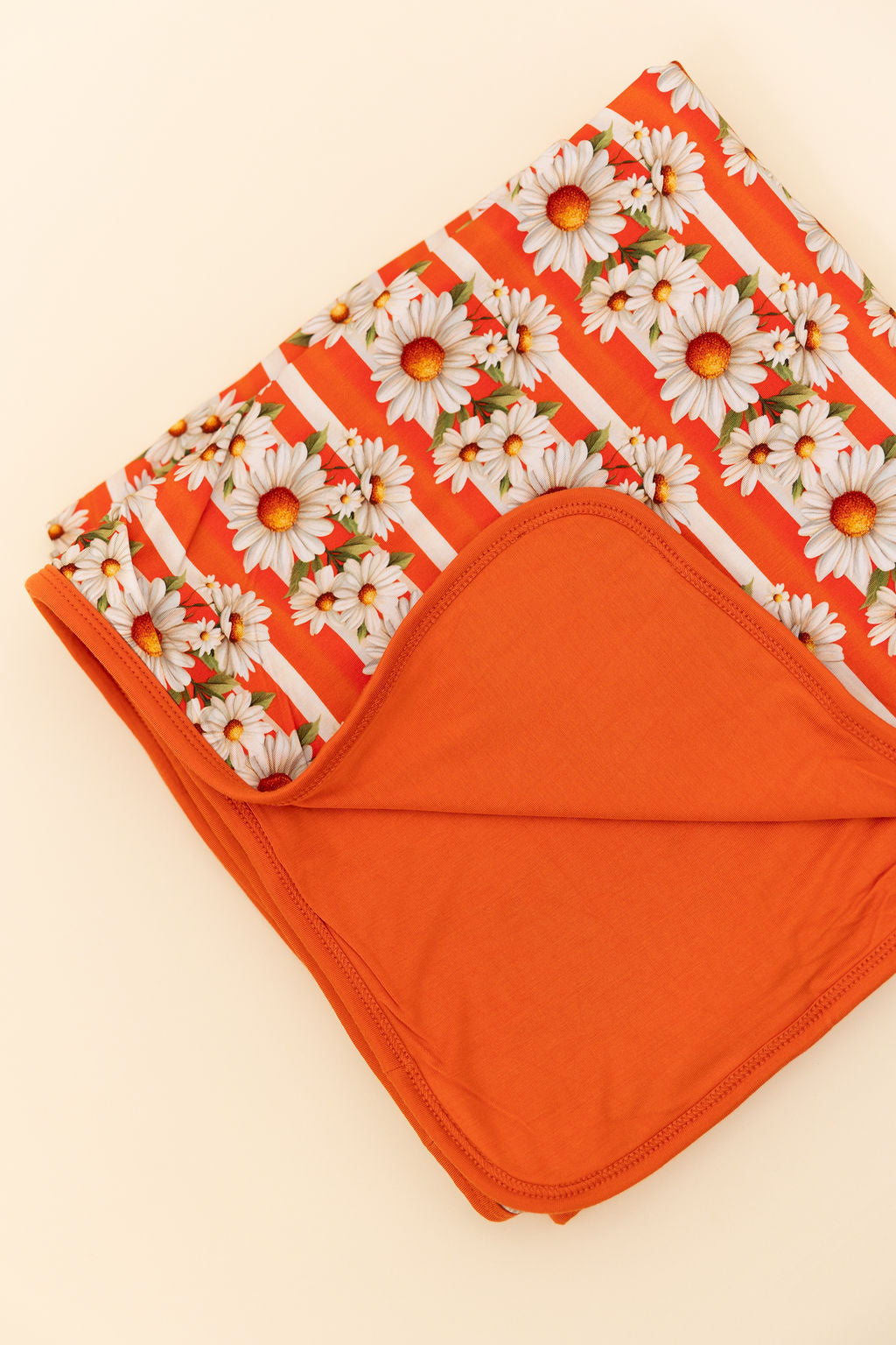 Close up of a bamboo reversible blanket orange and white stripe, with white daisy's on top  and the reverse side a bright orange against a beige background