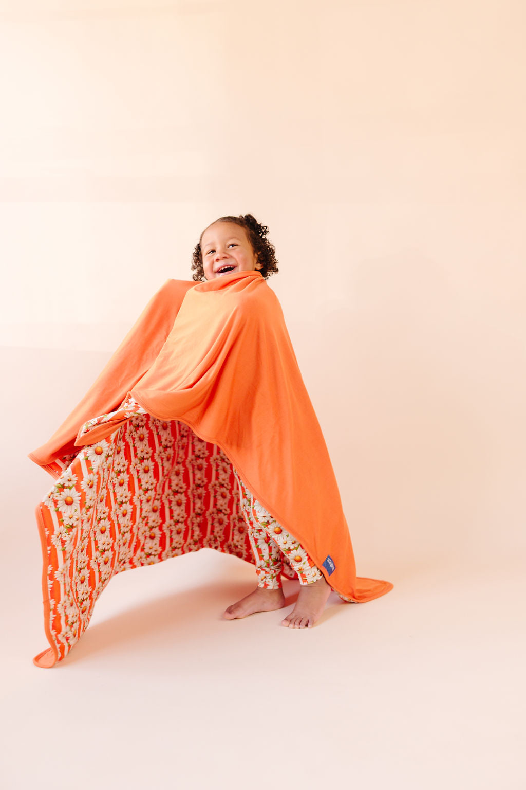 Girl spinning in a bamboo blanket, pattern on blanket is orange and white stripe, with white daisy's on top  and the reverse side a bright orange against a beige background