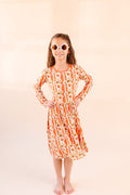 Young girl in flower sunglasses wearing a bamboo Twirl dress with orange and white stripe, with white daisy's on top  and the reverse side a bright orange against a beige background