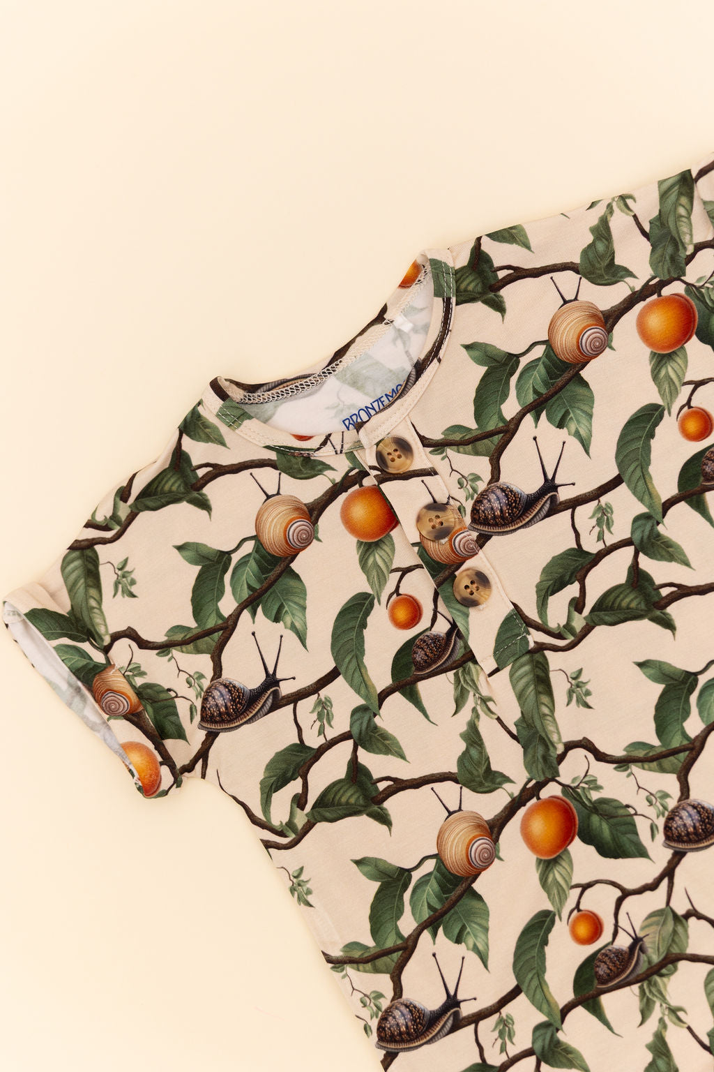 Close up flat lay image of Bamboo fabric Short sleeve baby Romper with a nature-themed pattern featuring snails, apricots, branches and leaves on a beige background.