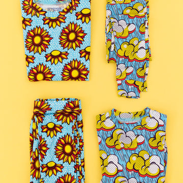Four folded pieces of bright pop art inspired clothing laid out on a bright yellow background, featuring blue, yellow, orange, and white designs including sunflowers and clouds.